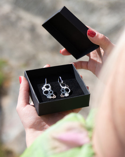 Rannalla-korumalliston kiiltävät hopeiset korvakorut koukkukiinnityksellä. Malli pitelee kuvassa korut sisältävää avonaista mustaa korurasiaa käsissään.