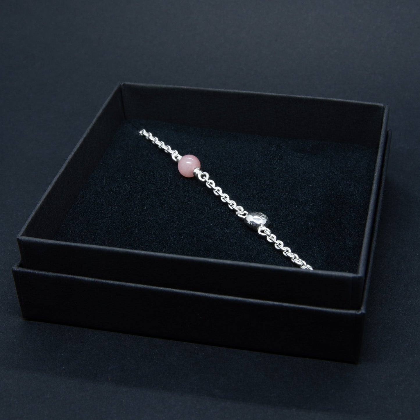 Hopeinen naisten rannekoru vaaleanpunaisella kivihelmellä ja käsityönä tehdyllä yksityiskohdalla. Koru on mustassa pahvisessa korurasiassa.