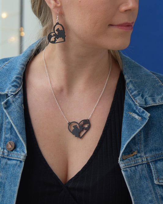 Oulu-aiheinen musta vanerikaulakoru, josta voi tunnistaa Toripoliisin ja Oulun Kauppahallin siluetit. Korussa on siro hopeinen papuketju.