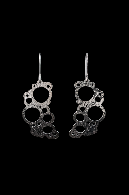 Hopeiset kiiltävät korvakorut koukkukiinnityksellä. Korujen pinta on kiiltäv, hieman kaareva ja vasaroitu pallopäävasaralla, joten se kiiltää eläväisesti. Korut ovat muotoilultaan orgaaniset ja ilmavat, sillä niissä on epäsäännöllinen kehämuoto ja useita pyöreitä läpimenoja.