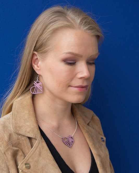 Rovaniemellä sijaistsevasta Jätkänkynttilä sillasta inspiroituneet mustikkamaidon sävyiset vanerikorvakorut ja kaulakoru vaaleahiuksisen mallin yllä. Korujen ulkokehä on sydämen mallinen.