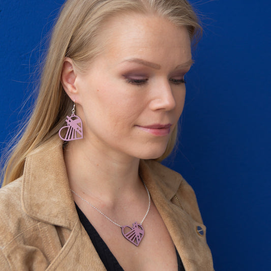 Rovaniemellä sijaistsevasta Jätkänkynttilä sillasta inspiroituneet mustikkamaidon sävyiset vanerikorvakorut ja kaulakoru vaaleahiuksisen mallin yllä. Korujen ulkokehä on sydämen mallinen.