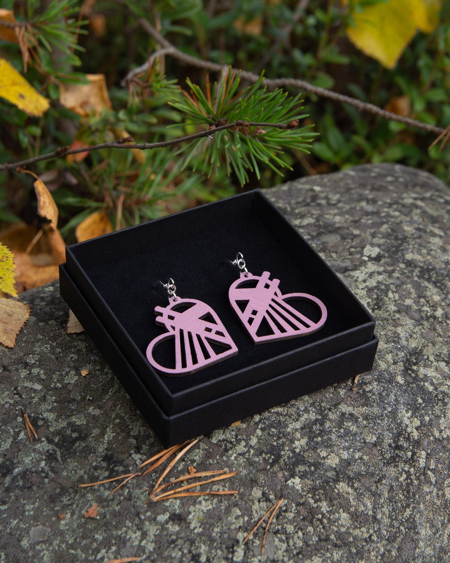 Vaaleanpunaisen sävyiset Rovaniemi-aiheiset vanerikorvakorut mustassa korurasiassa.